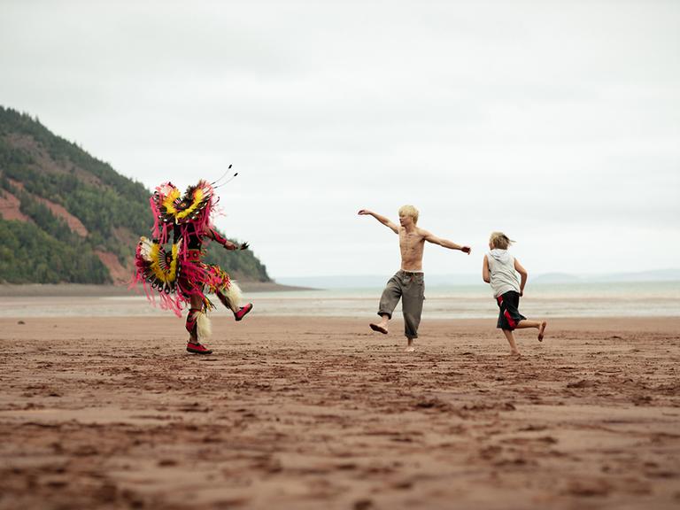 Eine Filmszene aus "Wildhood" zeigt drei Personen an einem sonst menschenleeren Meeresstrand. Zu sehen sind ein Mann in indigener Kostümierung und zwei Jungen beim Tanzen.