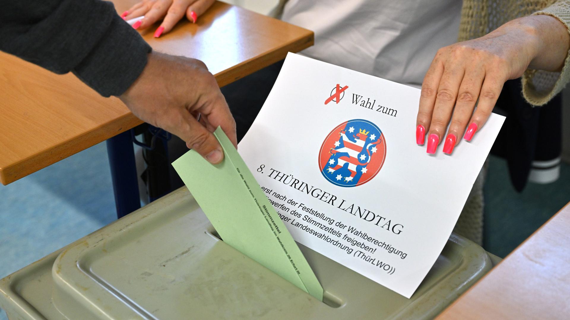 Die Hand eines Wählers steckt einen Stimmzettel in den Schlitz der grünen Wahlurne. Die Hand einer Wahlhelferin mit pinken langen Fingernägeln legt dafür die Abdeckung mit dem Schriftzug der Wahl zur Seite. 