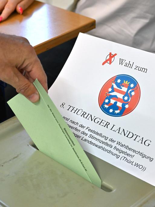 Die Hand eines Wählers steckt einen Stimmzettel in den Schlitz der grünen Wahlurne. Die Hand einer Wahlhelferin mit pinken langen Fingernägeln legt dafür die Abdeckung mit dem Schriftzug der Wahl zur Seite. 