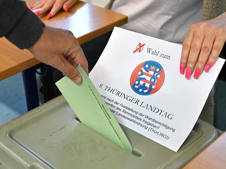 Die Hand eines Wählers steckt einen Stimmzettel in den Schlitz der grünen Wahlurne. Die Hand einer Wahlhelferin mit pinken langen Fingernägeln legt dafür die Abdeckung mit dem Schriftzug der Wahl zur Seite. 