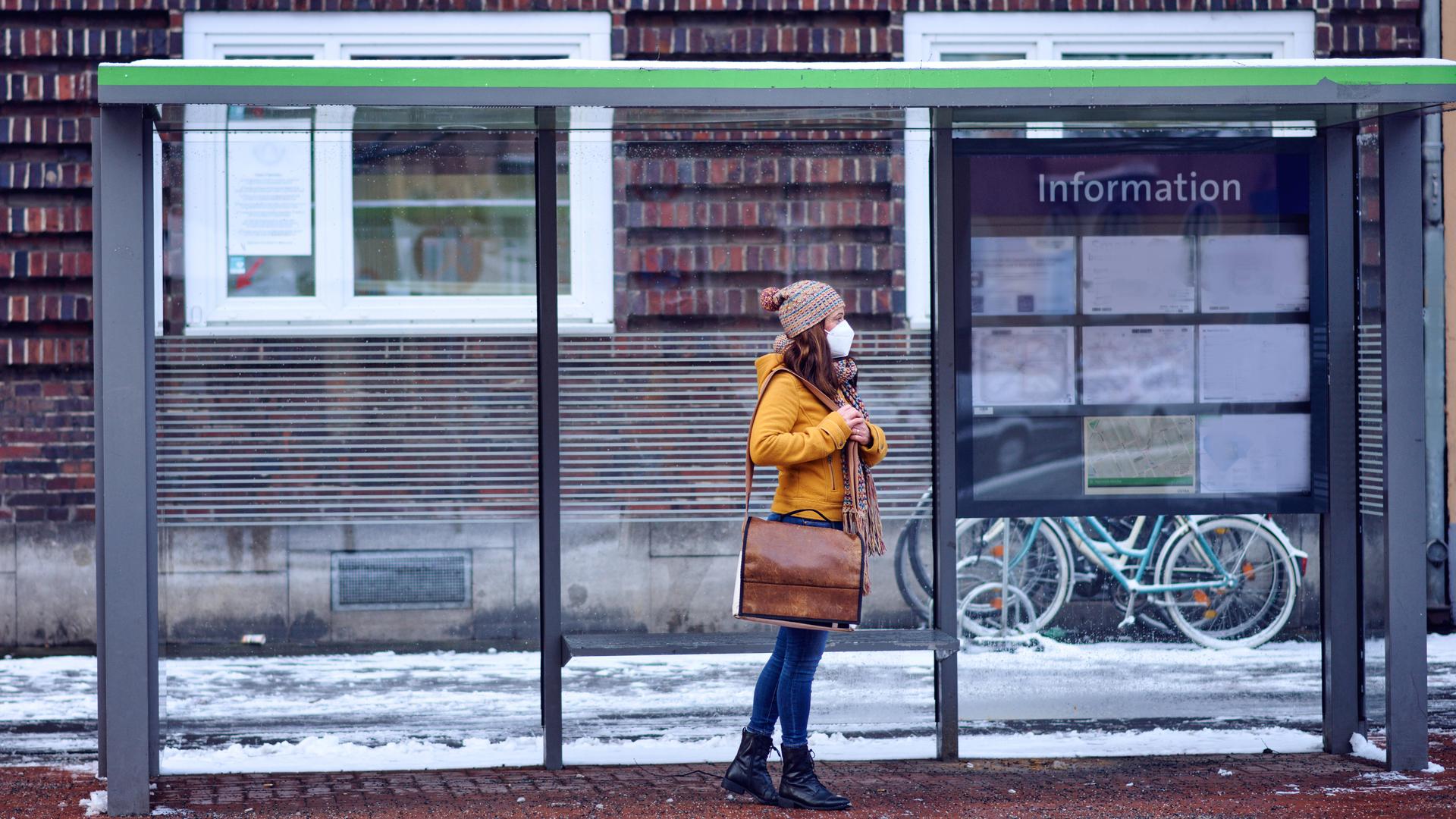 Eine Frau mit Mund-Nasen-Schutz steht an einer Bushaltestelle im Schnee.