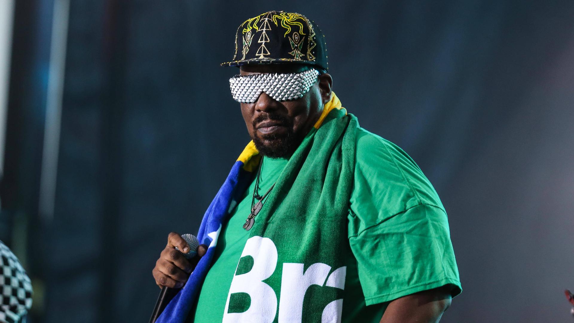 Afrika Bambaata hält ein Mikrofon. Er trägt eine Schirmmütze, ein grünes T-Shirt und eine silberne Sonnenbrille.
