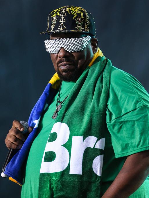 Afrika Bambaata hält ein Mikrofon. Er trägt eine Schirmmütze, ein grünes T-Shirt und eine silberne Sonnenbrille.