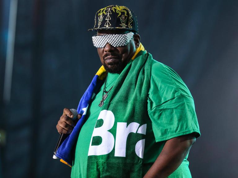 Afrika Bambaata hält ein Mikrofon. Er trägt eine Schirmmütze, ein grünes T-Shirt und eine silberne Sonnenbrille.