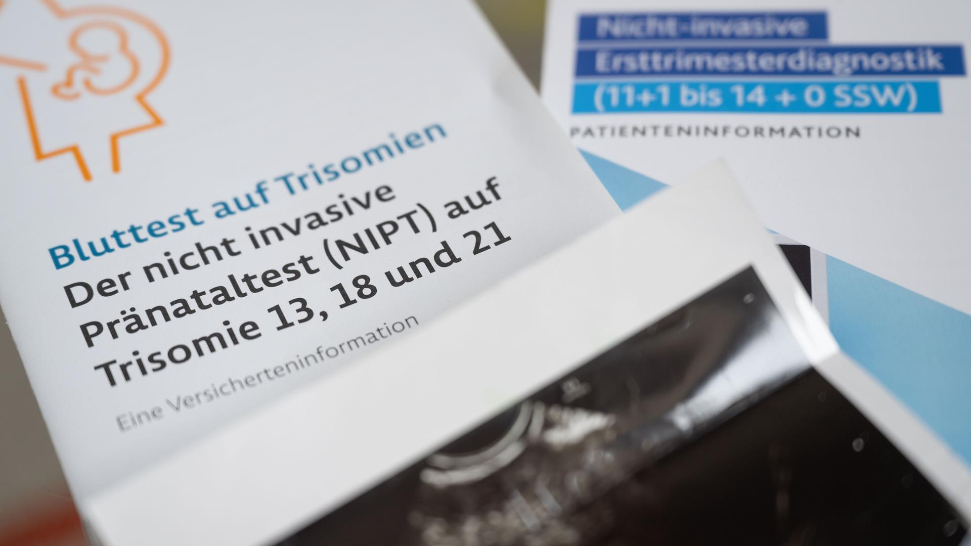 Zwei Informationsbroschüren zu nichtinvasiven Pränataltests liegen neben einem Ultraschallbild in einer Frauenarztpraxis.