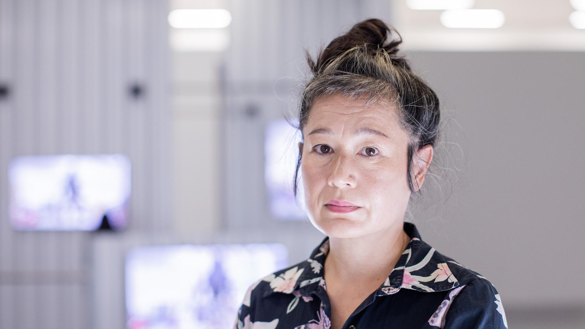 Portrait von Hito Steyerl, die in der Kunstsammlung K21 steht. Im Hintergrund sind im Raum aufgehängte Monitore zu sehen.