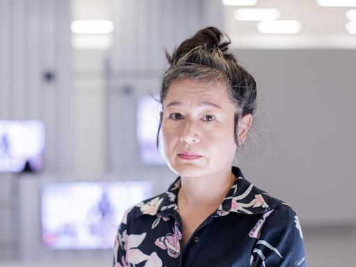 Portrait von Hito Steyerl, die in der Kunstsammlung K21 steht. Im Hintergrund sind im Raum aufgehängte Monitore zu sehen.