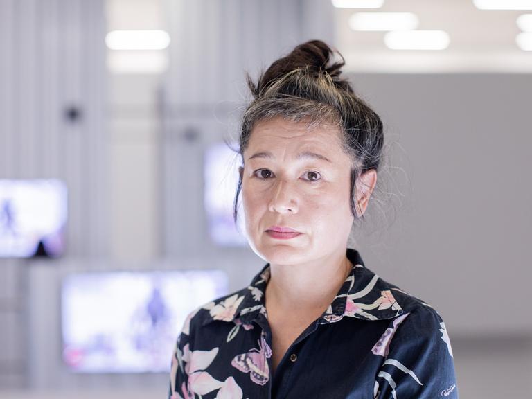 Portrait von Hito Steyerl, die in der Kunstsammlung K21 steht. Im Hintergrund sind im Raum aufgehängte Monitore zu sehen.