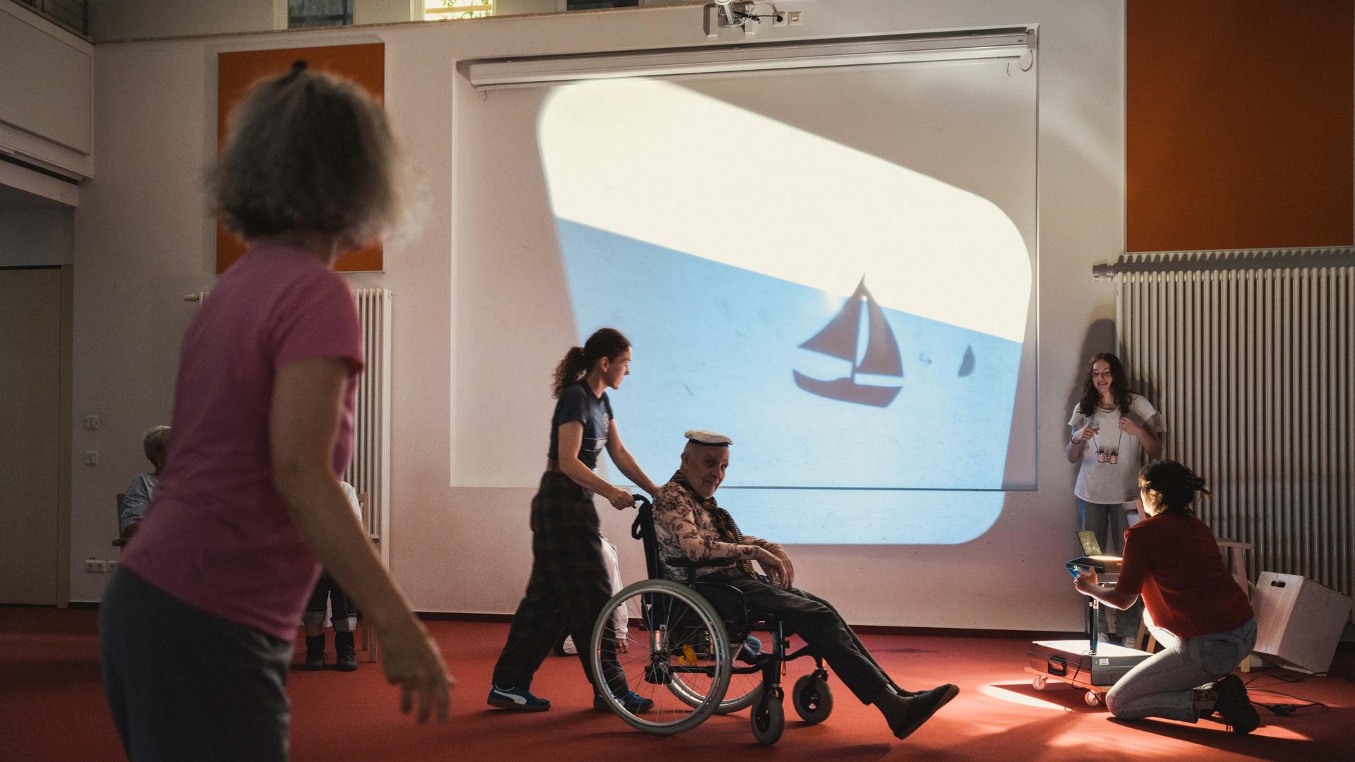 Darsteller der Theatergruppe "Papillon" spielen auf einer Bühne vor einem Hintergrund mit Schiff, ein Schauspieler wird im Rollstuhl gefahren.