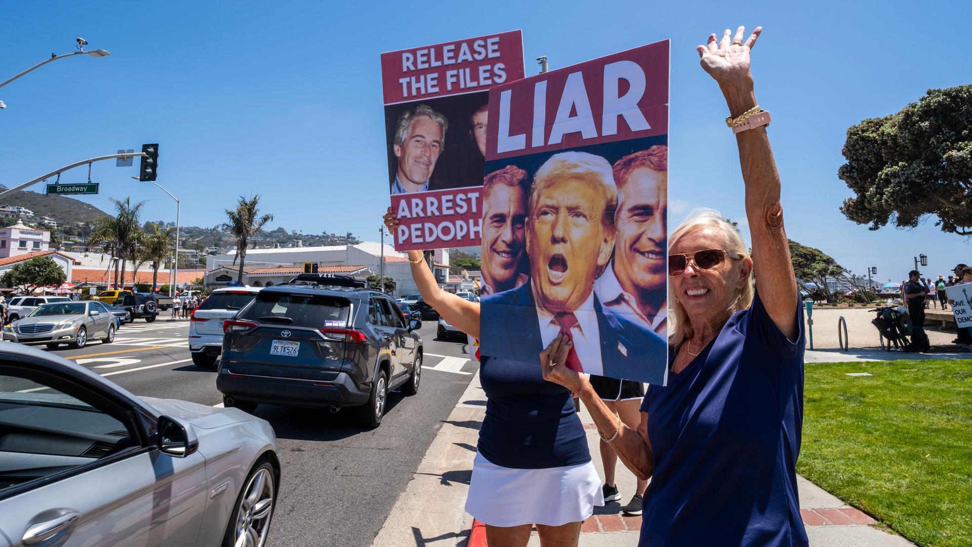 Zwei Demonstrantinnen halten Plakate hoch, auf denen die Köpfe von Jeffrey Epstein und Donald Trump zu sehen sind. Zu lesen ist "Release the Files", "Arrest Pedophiles" und "Liar".