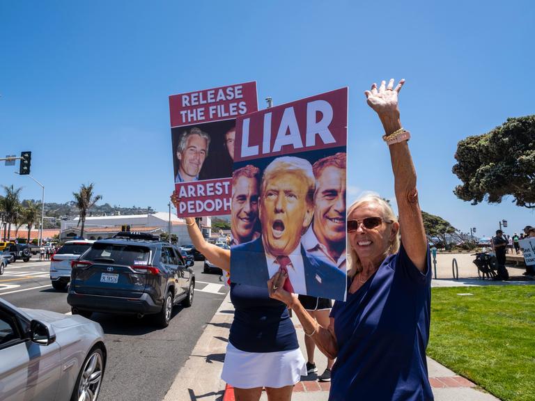 Zwei Demonstrantinnen halten Plakate hoch, auf denen die Köpfe von Jeffrey Epstein und Donald Trump zu sehen sind. Zu lesen ist "Release the Files", "Arrest Pedophiles" und "Liar".