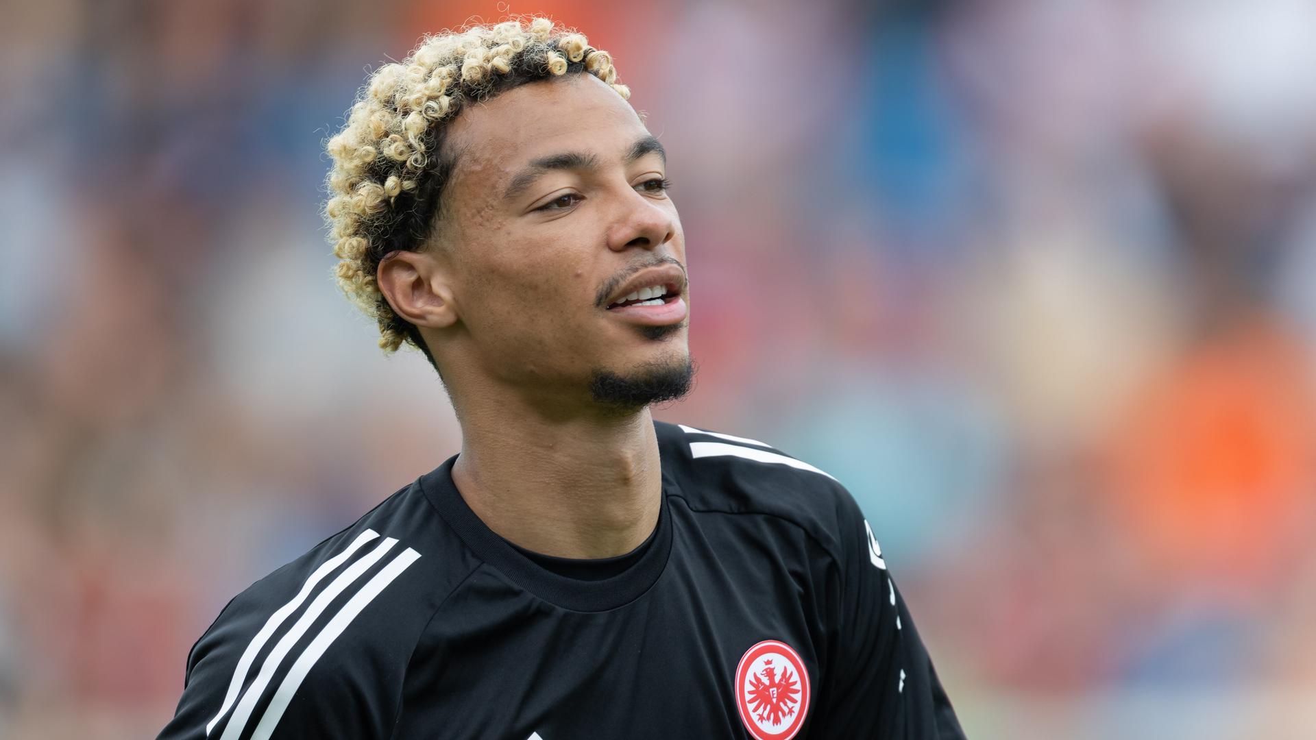 Portrait von einem jungen, schwarzen Mann mit blond gefärbten Locken im schwarzen Eintracht-Frankfurt-Trikot.
