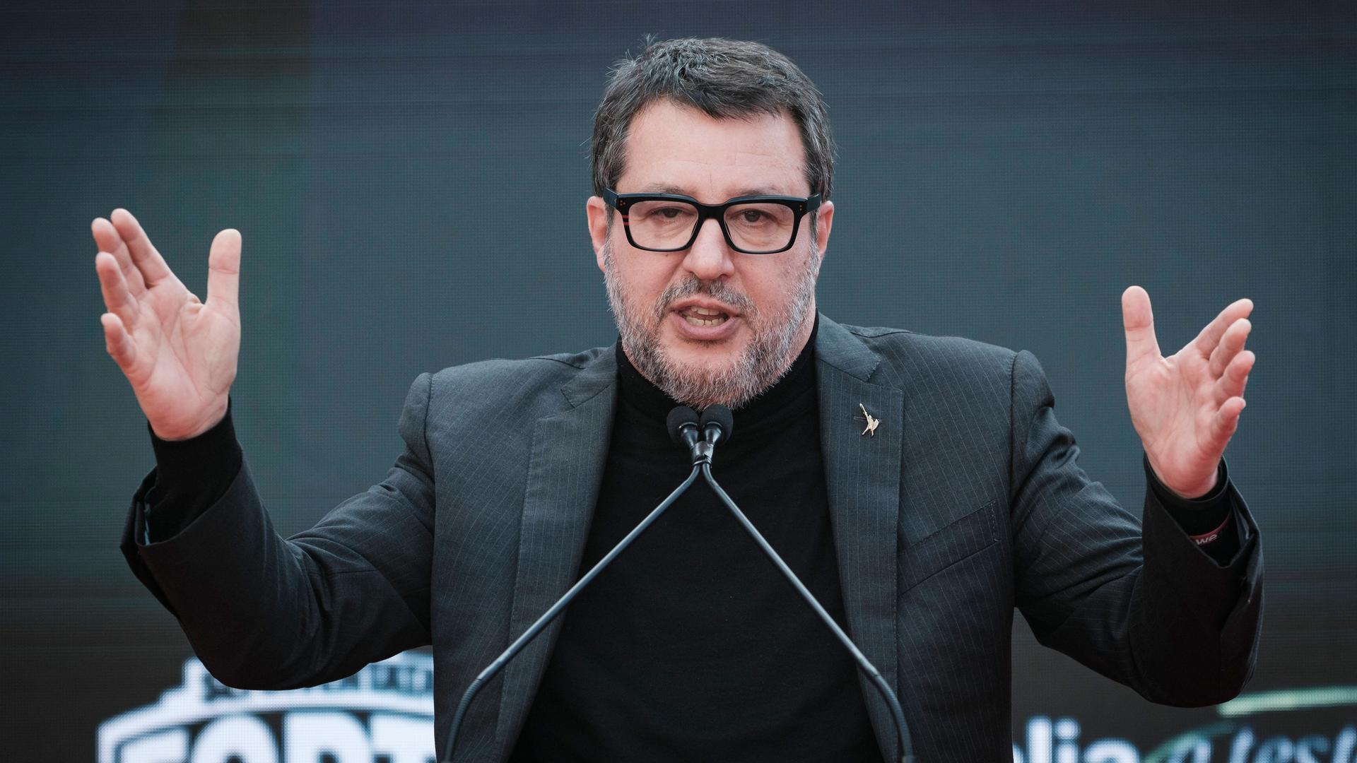 Ein mittelalter Mann mit grauem Bart und Haaren sowie Brille. Er trägt einen dunklen Rollkragenpullover und Jackett und steht vor einem Mikrofon und hebt beide Arme. Ein mittelalter Mann mit grauem Bart und Haaren sowie Brille. Er trägt einen dunklen Rollkragenpullover und Jackett und steht vor einem Mikrofon und hebt beide Arme.