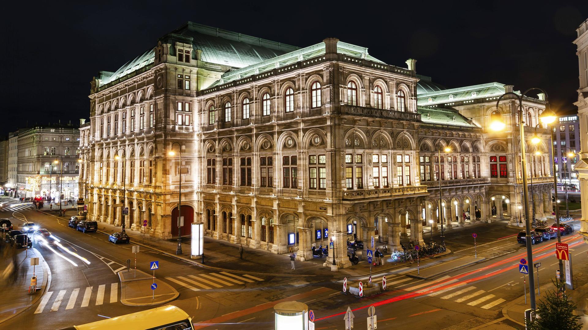 Die Wiener Staatsoper mit Straßenverkehr bei Nacht. Das Gebäude ist im repräsentativen Stil des 19. Jahrhunderts gebaut.