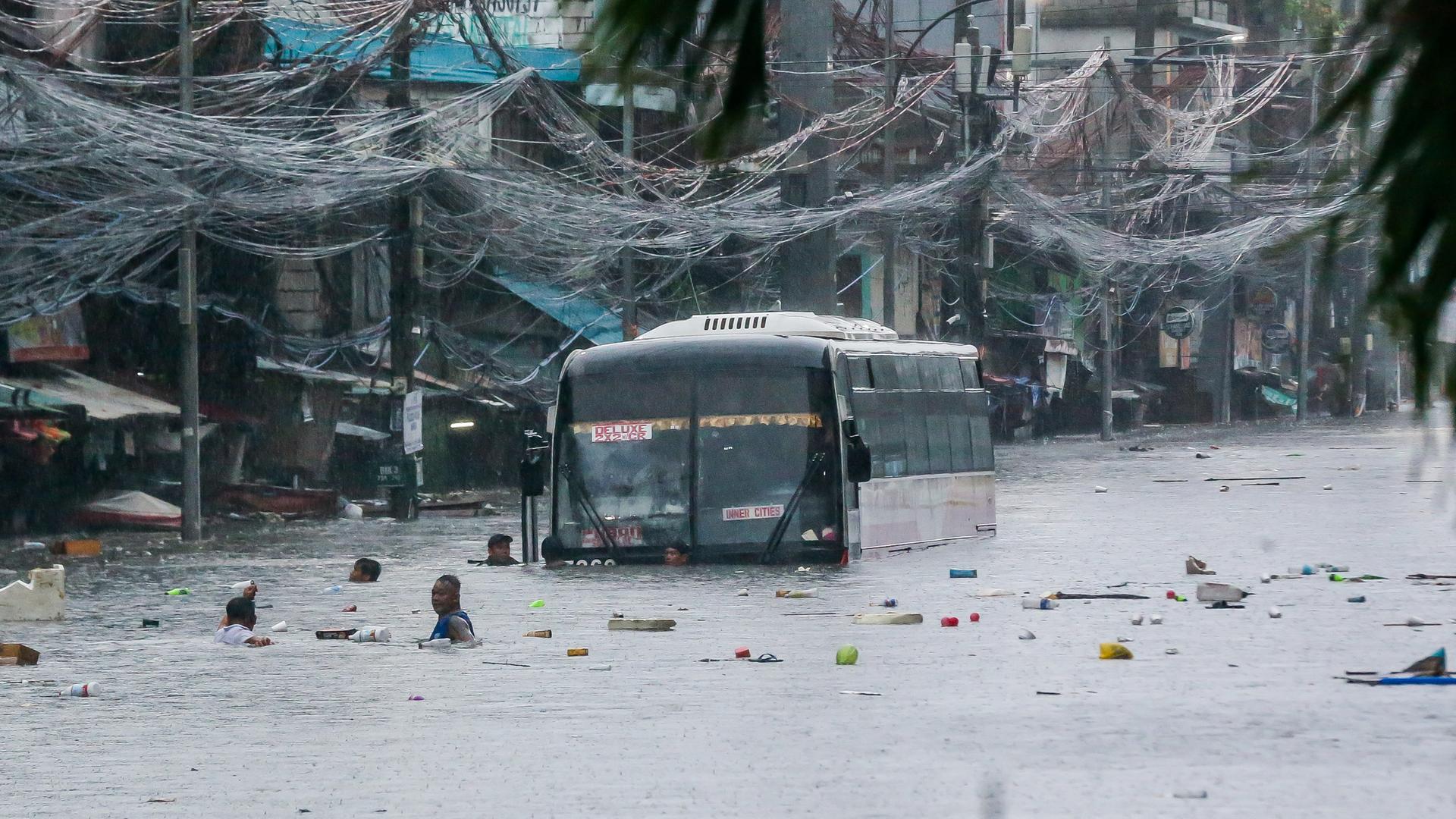 Blick aus der Ferne auf den Bus, der bis zum unteren Rand der Frontscheibe in einer Straße mit Häusern im Wasser steht. Mehrere Menschen stehen bis zum Kopf in den Fluten, Müll schwimmt darauf. 