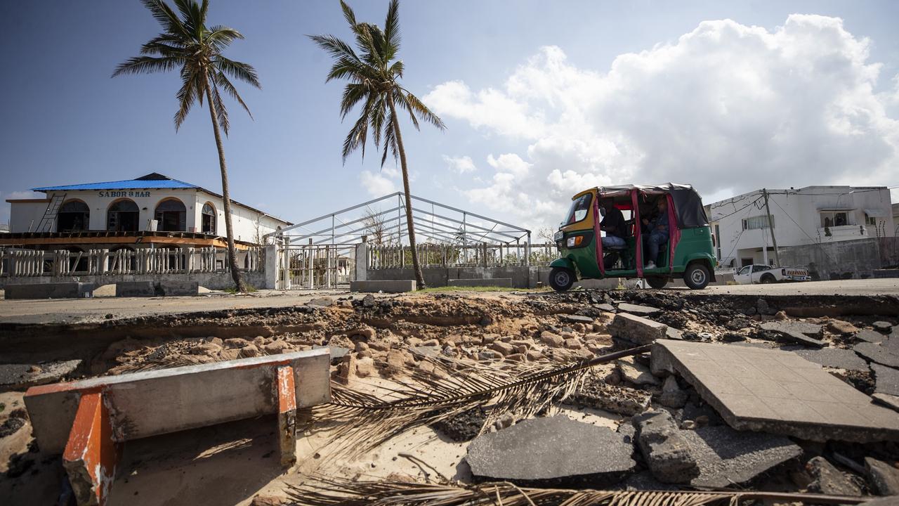 Trümmer liegen vor einer Straße nach einem Zyklon, auf der ein Tuktuk unterwegs ist; im Hintergrund Palmen, abgedeckte Häuser und blauer Himmel.