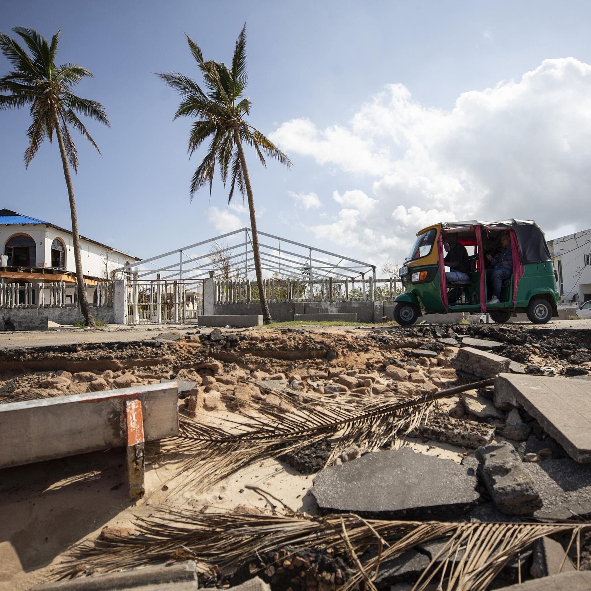 Trümmer liegen vor einer Straße, auf der ein Tuktuk unterwegs ist; im Hintergrund Palmen, abgedeckte Häuser und blauer Himmel.