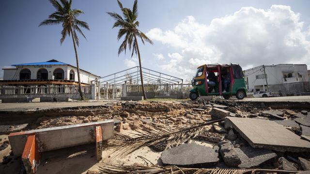 Trümmer liegen vor einer Straße, auf der ein Tuktuk unterwegs ist; im Hintergrund Palmen, abgedeckte Häuser und blauer Himmel.