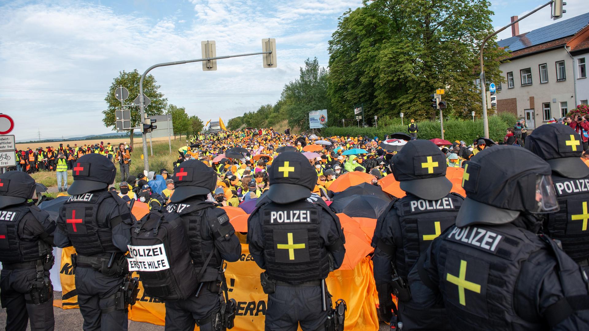 Auf einer Landstraße sitzt eine große Menschenmenge, davor steht eine Reihe von Polizisten. Auf einer Landstraße sitzt eine große Menschenmenge, davor steht eine Reihe von Polizisten.