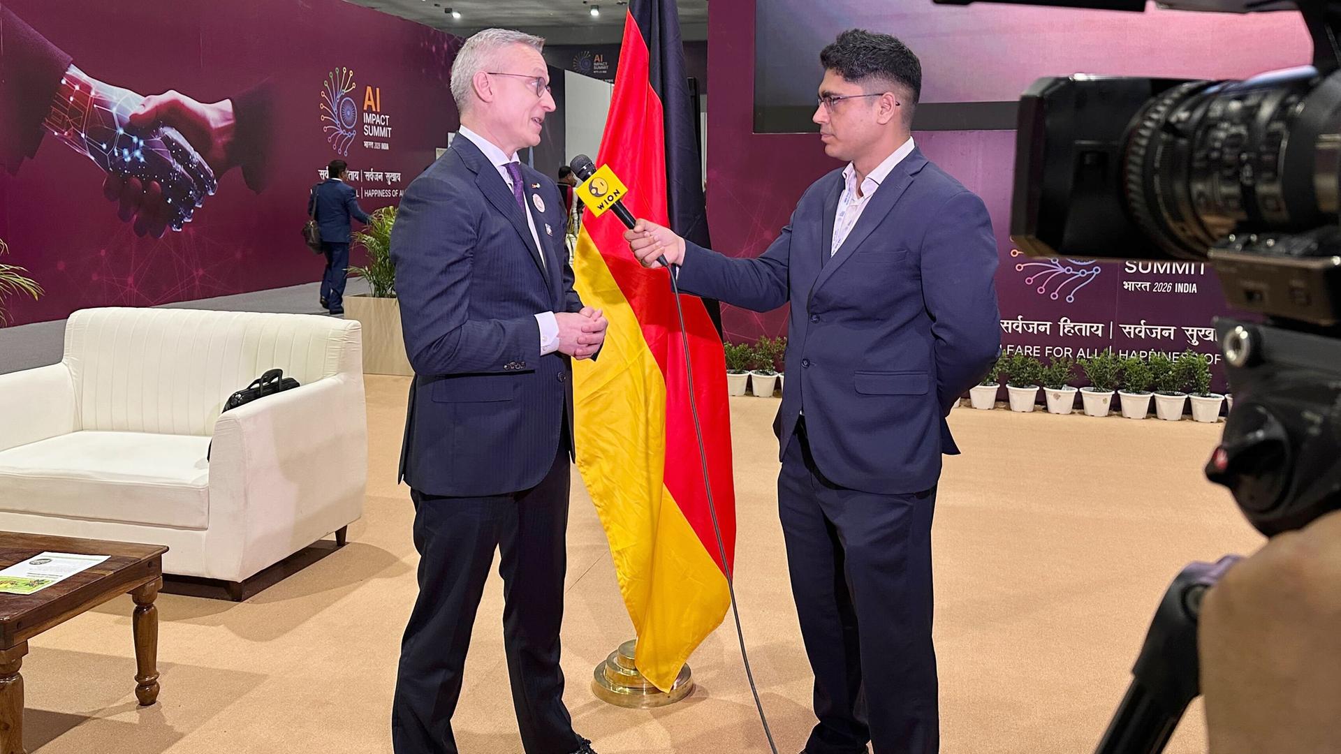 Bundesdigitalminister Karsten Wildberger spricht in ein Mikrofon, das ihm ein anderer Mann hinhält. Neben ihnen ist eine Deutschlandflagge.