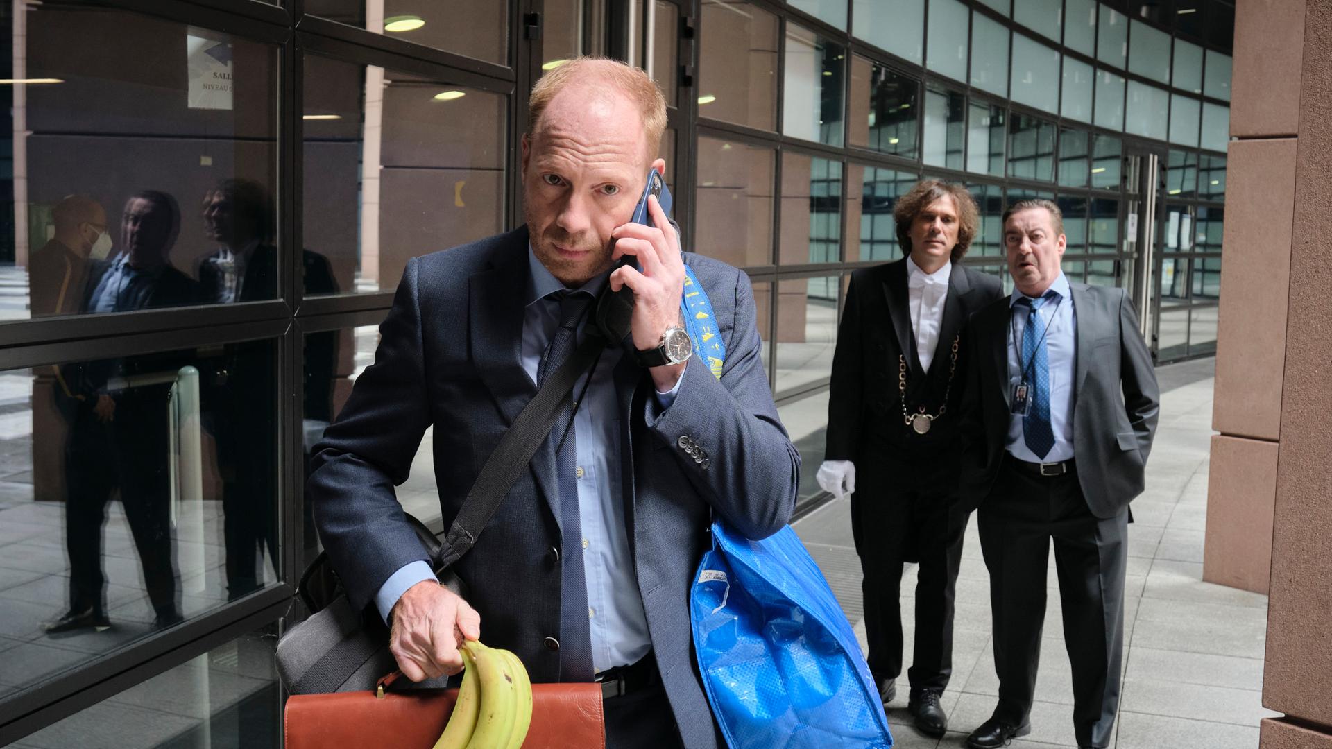 Ein Szenenfoto aus der ARD-/WDR-Serie "Parlament": Der Schauspieler Johann von Bülow steht im Vordergrund und telefoniert mit seinem Smartphone, während er eine Bürotasche hält und eine blaue IKEA-Tasche über der Schulter trägt, Seine rechte Hand hält zudem ein Bündel mit Bananen. Im Hintergrund stehen zwei Männer und beobachten ihn. 