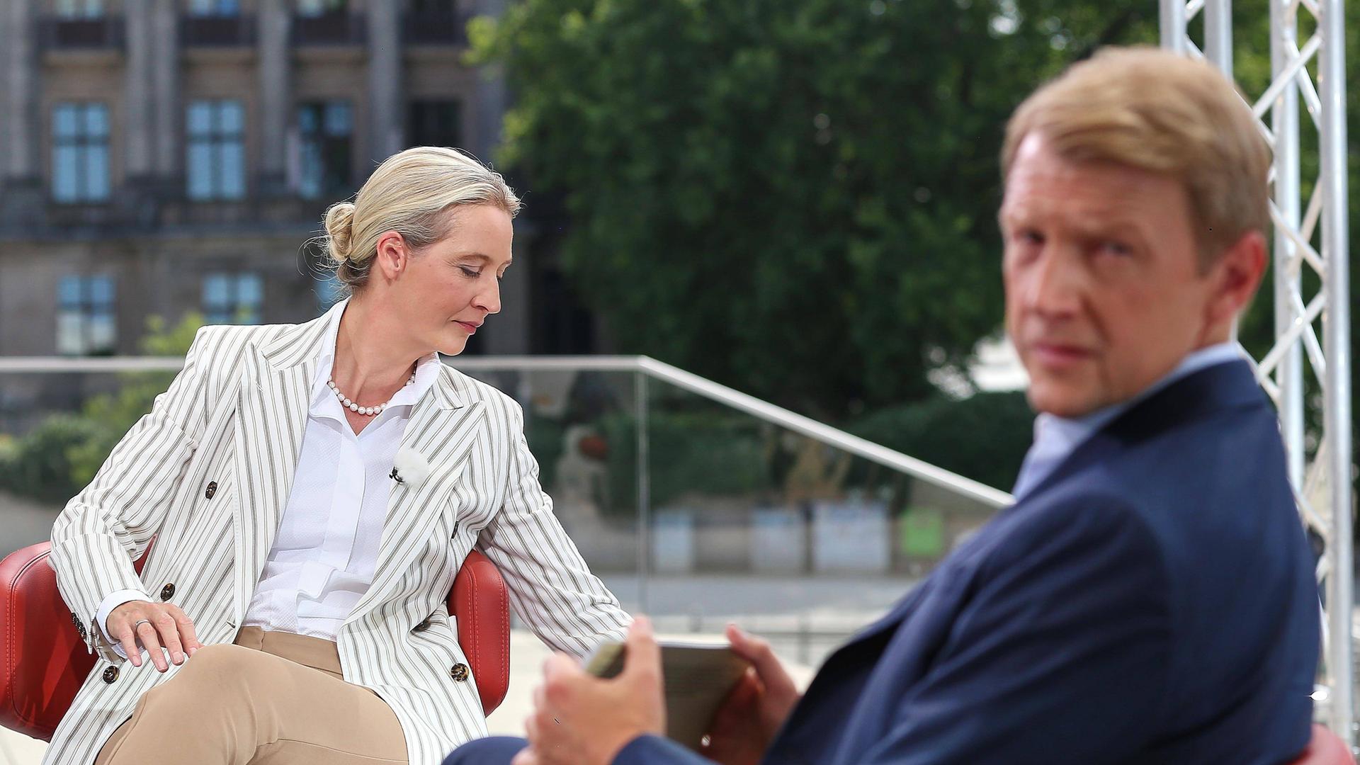 AfD-Vorsitzende Alice Weidel sitzt in einem hellen Anzug auf einem roten Sessel und dreht sich zur Seite. ARD-Moderator Markus Preiß schaut über seine Schulter in Richtung Kamera. 