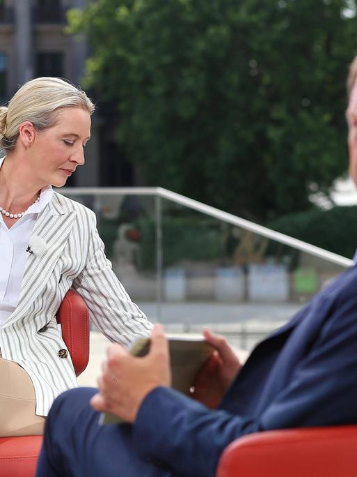 AfD-Vorsitzende Alice Weidel sitzt in einem hellen Anzug auf einem roten Sessel und dreht sich zur Seite. ARD-Moderator Markus Preiß schaut über seine Schulter in Richtung Kamera. Sein Gesicht ist verschwommen dargestellt. 