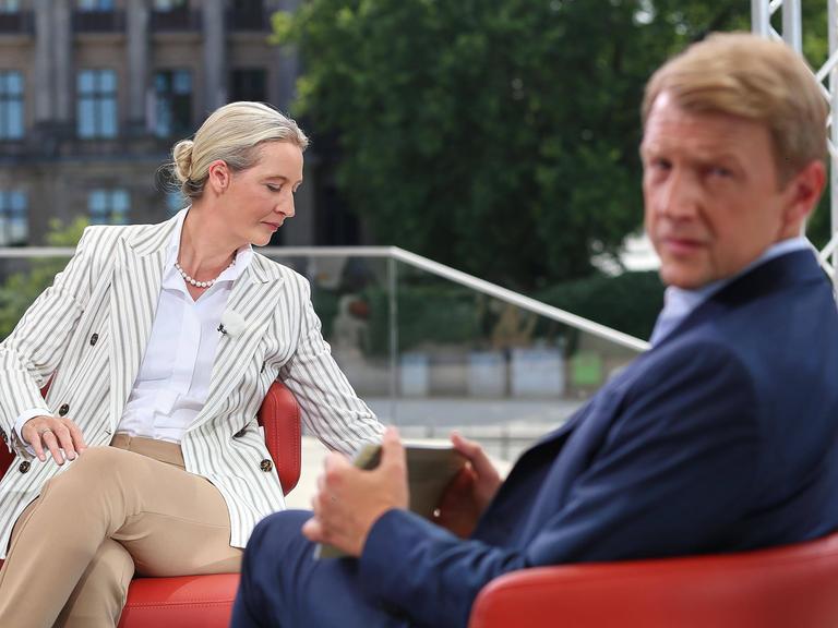 AfD-Vorsitzende Alice Weidel sitzt in einem hellen Anzug auf einem roten Sessel und dreht sich zur Seite. ARD-Moderator Markus Preiß schaut über seine Schulter in Richtung Kamera. Sein Gesicht ist verschwommen dargestellt. 