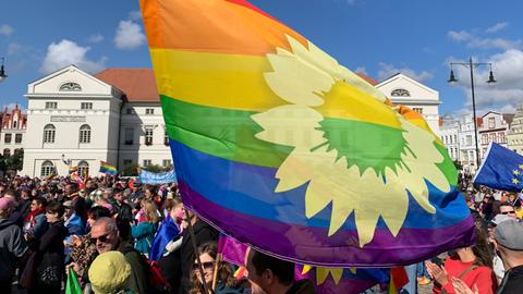 Teilnehmer stehen zusammen. Im Vordergrund schwenkt jemand eine große Regenbogenfahne.