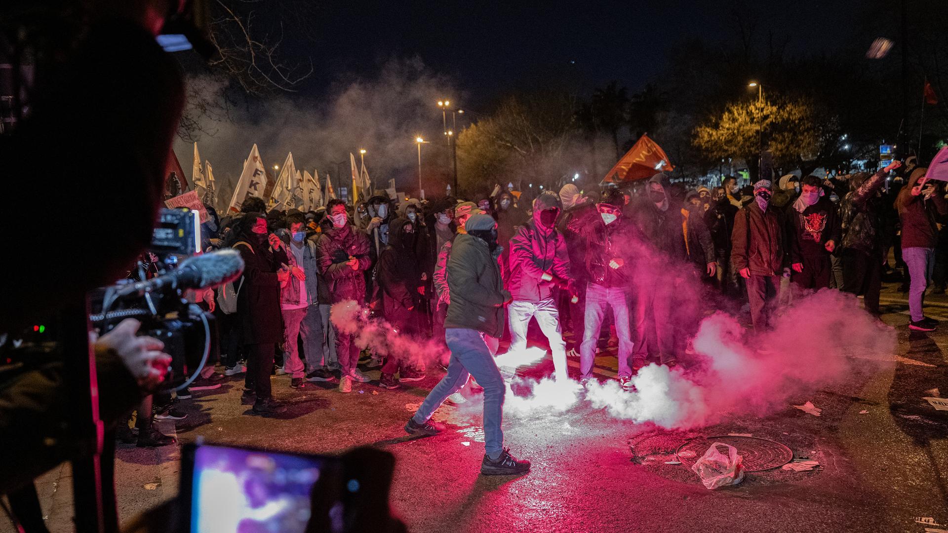 Auf einer Straße haben sich teils vermummte Menschen versammelt. Einige zünden Pyrotechnik. Kameraleute filmen die Szene.