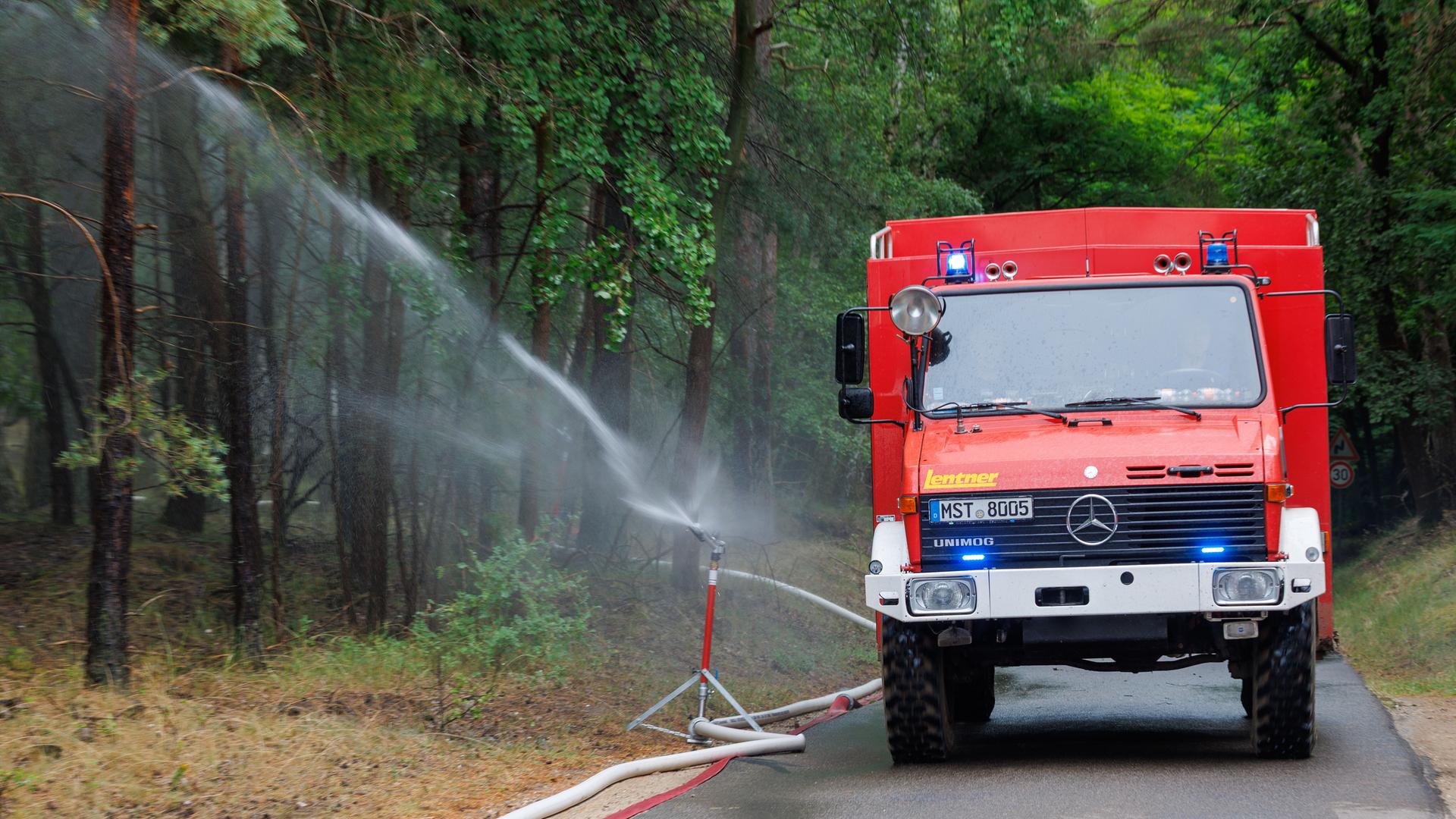 Das Bild zeigt ein Auto von der Feuer-Wehr. Es steht auf einer Straße. Neben dem Auto steht ein Wasser-Sprinkler. Der Wasser-Sprinkler spritzt Wasser auf die Bäume. Das Bild zeigt ein Auto von der Feuer-Wehr. Es steht auf einer Straße. Neben dem Auto steht ein Wasser-Sprinkler. Der Wasser-Sprinkler spritzt Wasser auf die Bäume.