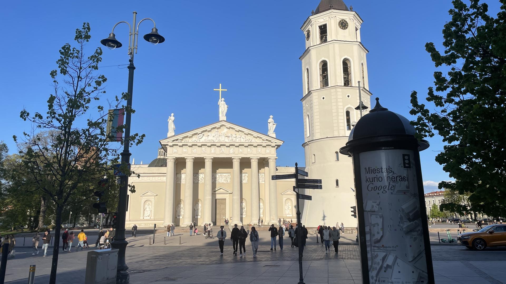 Alltag an einem Frühlingstag in Litauens Hauptstadt Vilnius. Menschen gehen spazieren, im Hintergrund die Kathedrale der Stadt, rechts im Vordergrund eine Litfaßsäule.