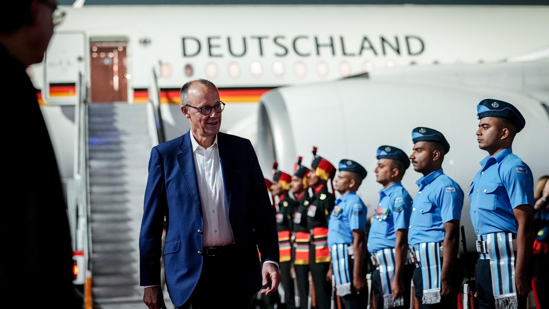 Merz läuft lächelnd an einer Reihe strammstehender indischer Soldaten in hellblauen Paradeuniformen vorbei. Im Hintergrund die weiße Regierungsmaschine und die Gangway. 