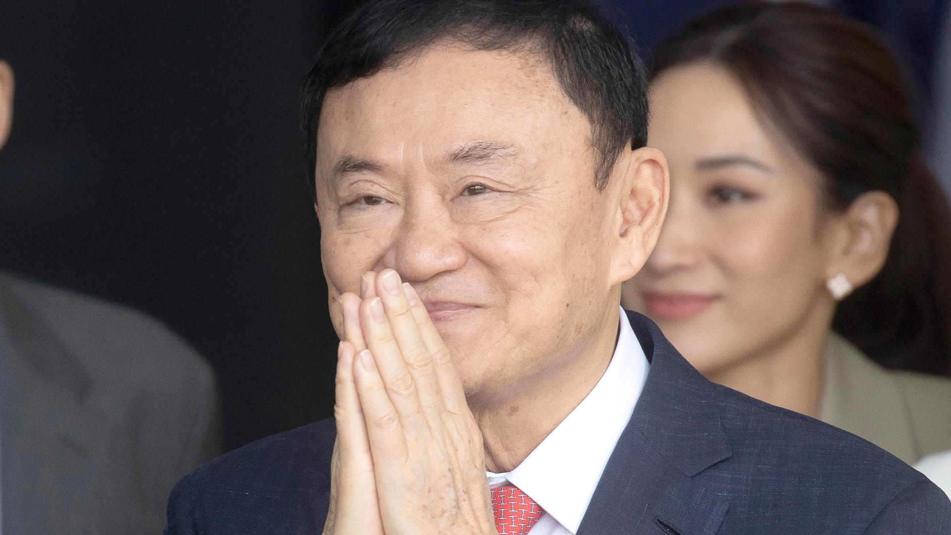 Thaksin faltet vor seinem Gesicht lächelnd die Hände. Dahinter eine ebenfalls lächelende Frau.