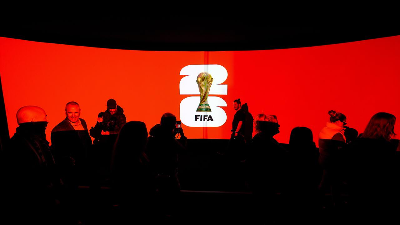 Das Logo der WM 2026 vor einem roten Hintergrund, daneben stehen Menschen.