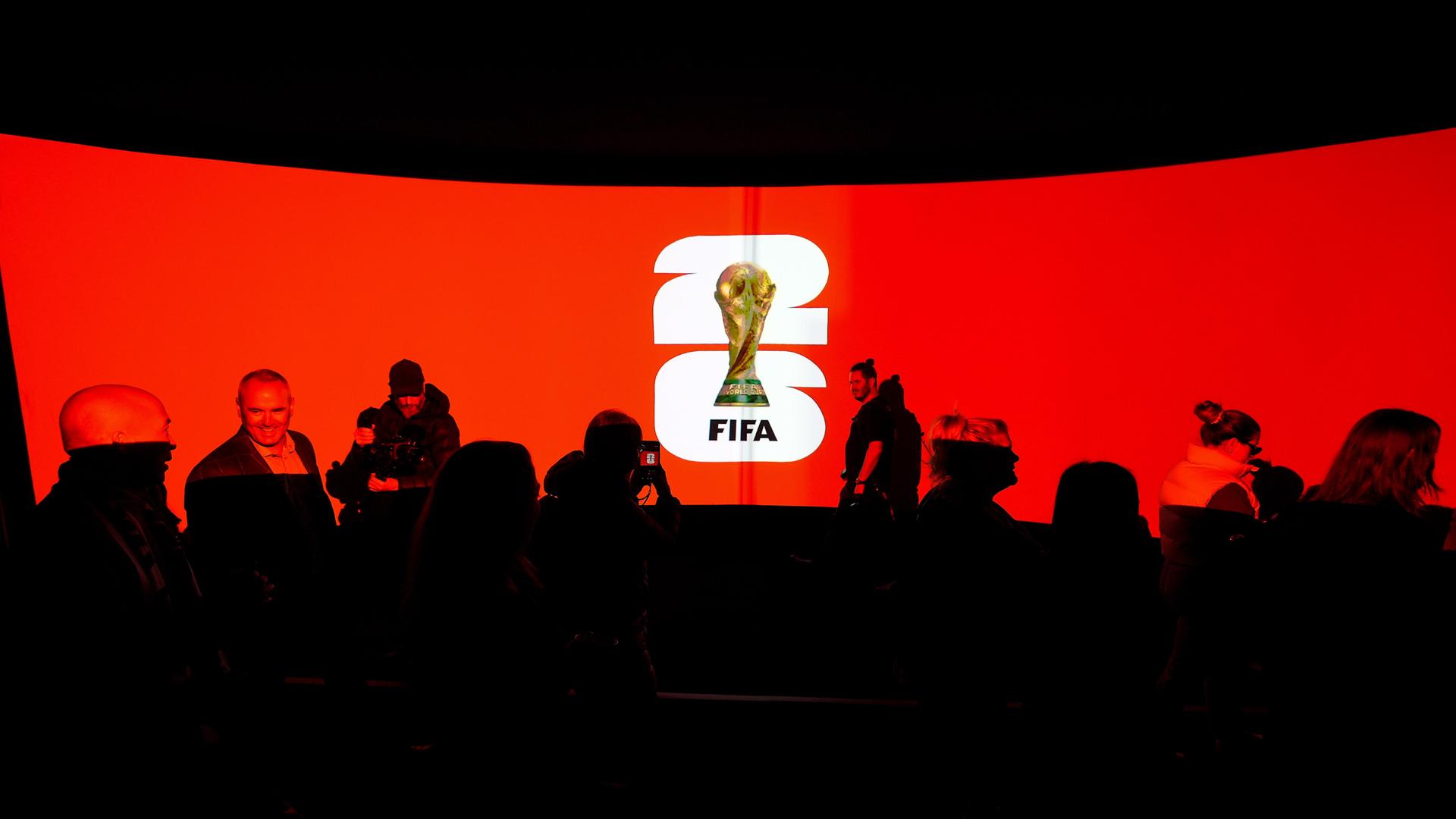 Das Logo der WM 2026 vor einem roten Hintergrund, daneben stehen Menschen.