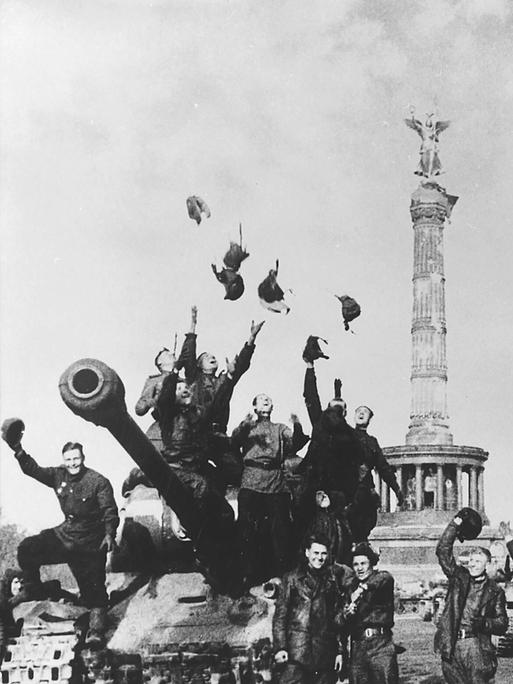 Historische Schwarz-Weiß-Aufnahme von sowjetischen Soldaten, die vor der Siegessäule in Berlin jubelnd ihre Helme und Hüte in die Luft werfen.