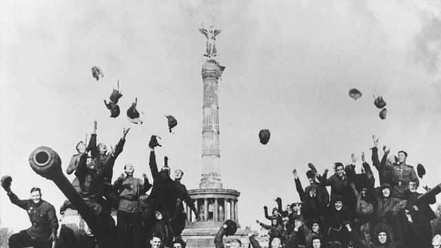 Historische Schwarz-Weiß-Aufnahme von sowjetischen Soldaten, die vor der Siegessäule in Berlin jubelnd ihre Helme und Hüte in die Luft werfen.