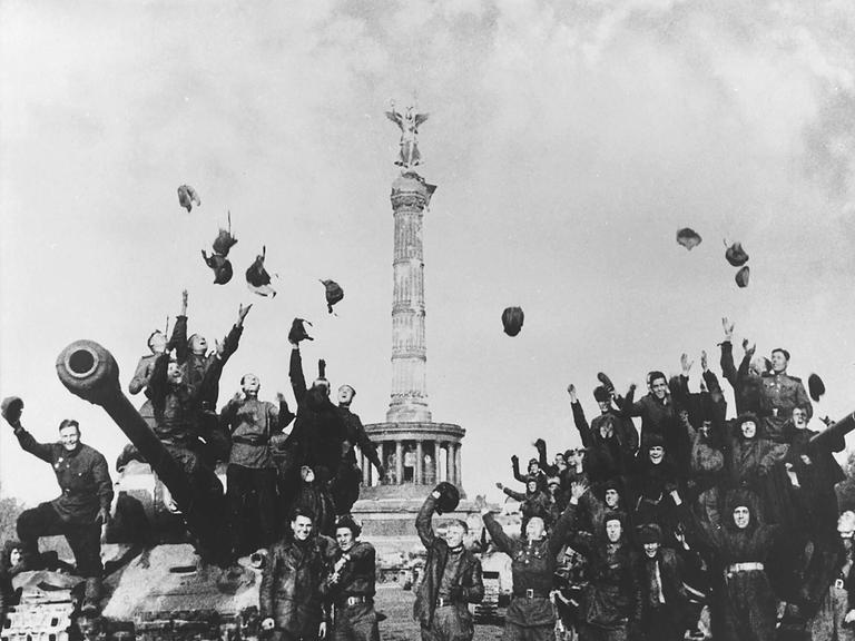 Historische Schwarz-Weiß-Aufnahme von sowjetischen Soldaten, die vor der Siegessäule in Berlin jubelnd ihre Helme und Hüte in die Luft werfen.