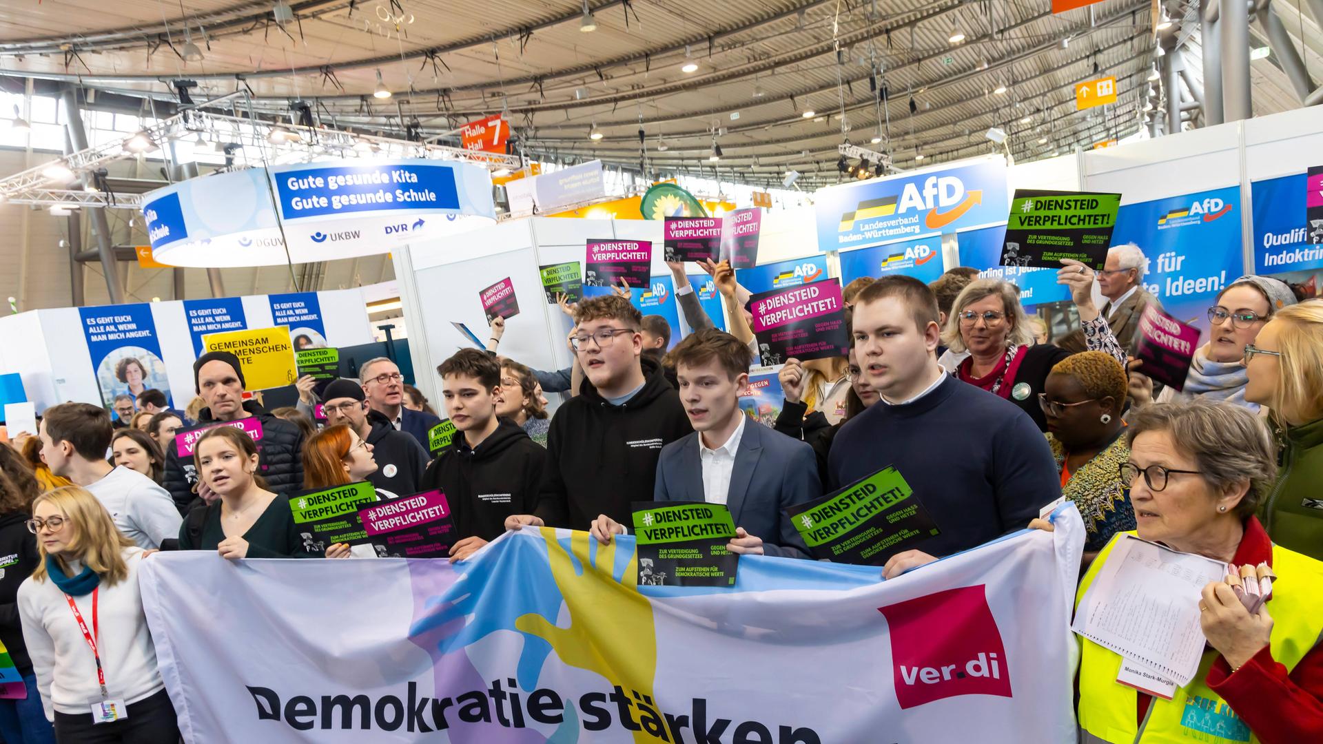 Das Foto zeigt mehrere Menschen, die gegen den AfD-Stand bei der Messe Didacta protestieren. Sie halten ein Banner mit der Aufschrift Demokratie stärken - Kampf gegen Rechts. 