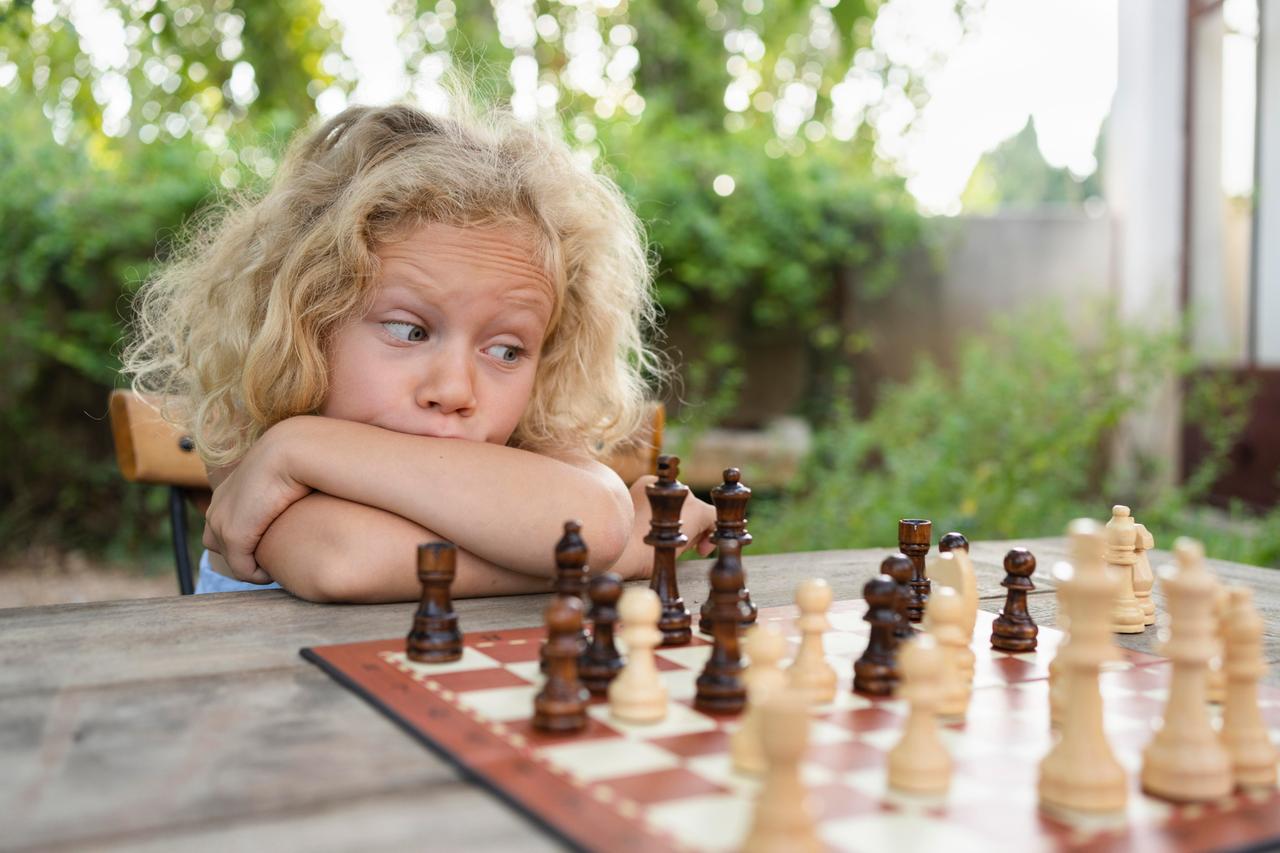 Ein am Tisch sitzendes Mädchen spielt eine Partie Schach und betrachtet das Feld nachdenklich.