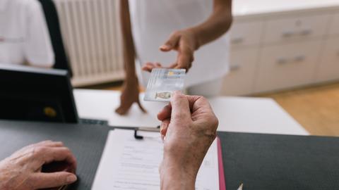 Eine Person reicht ihre Krankenkassenkarten über die Theke einer Arztpraxis.