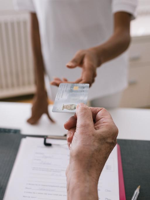 Eine Person reicht ihre Krankenkassenkarten über die Theke einer Arztpraxis.