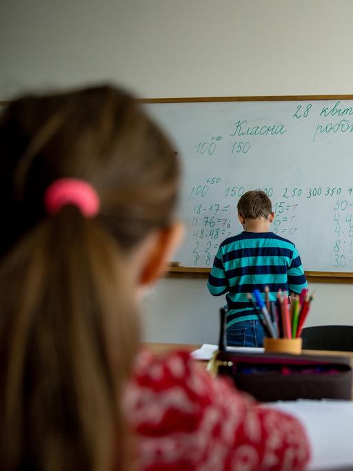 An einer Tafel die mit kyrillischen Buchstaben und mit Zahlen beschriftet sind, stehen zwei Kinder, die von hinten zu sehen sind. Im Vordergrund sind zwei weitere Kinder von hinten an einem Tisch zu sehen. 