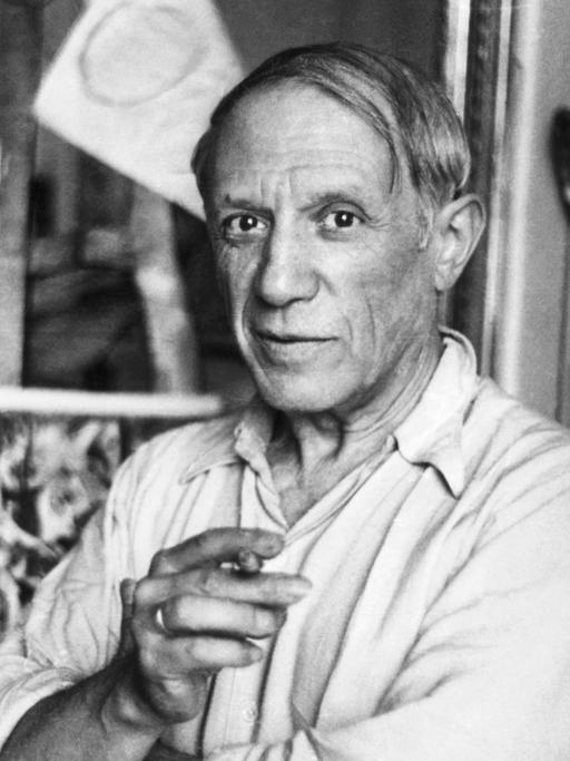 Pablo Picasso steht in seinem Studio, eine Zigarette in der Hand und schaut freundlich in die Kamera.