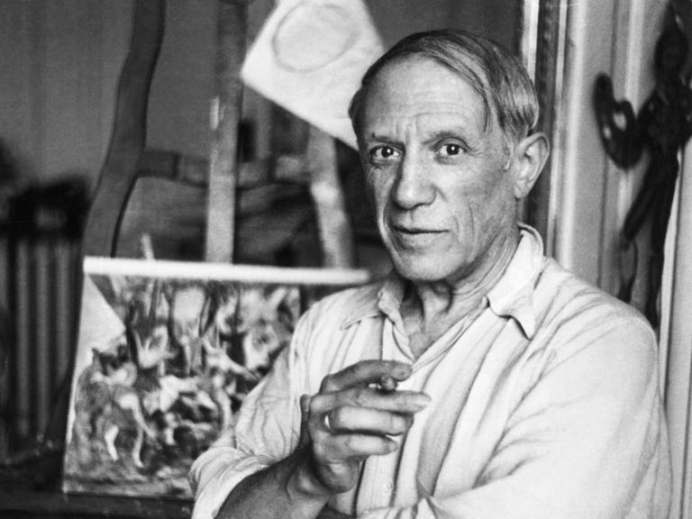 Pablo Picasso steht in seinem Studio, eine Zigarette in der Hand und schaut freundlich in die Kamera.