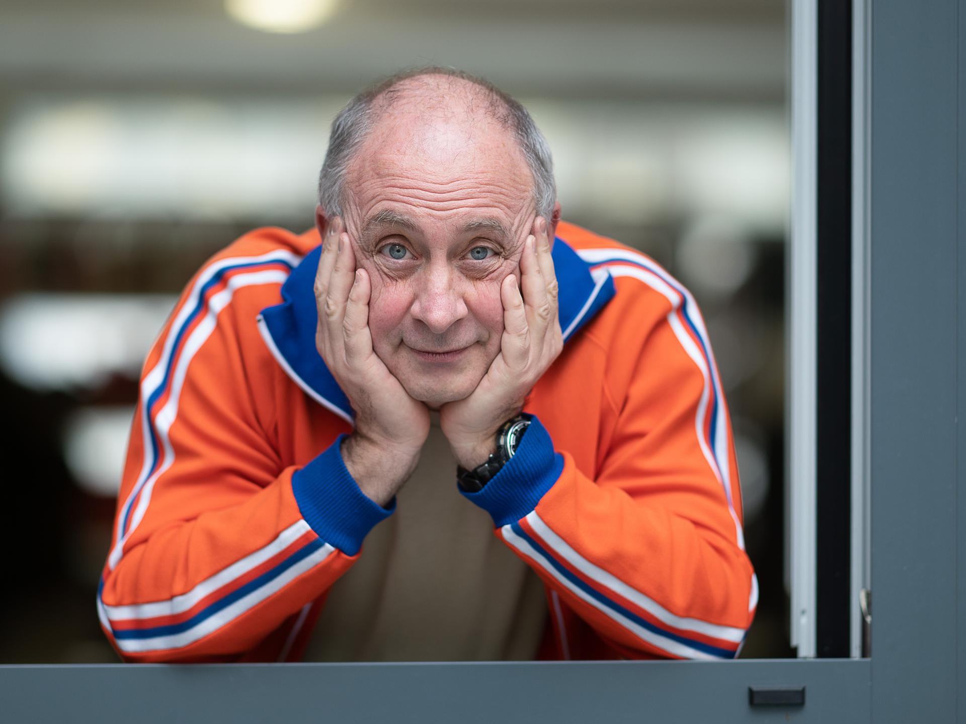 Alfons trägt eine orange-blaue Trainingsjacke und schaut, den Kopf auf die Hände gestützt, freundlich aus dem Fenster direkt in die Kamera.