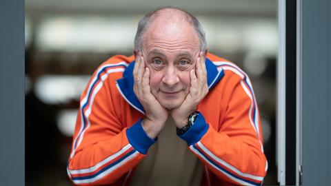 Alfons trägt eine orange-blaue Trainingsjacke und schaut, den Kopf auf die Hände gestützt, freundlich aus dem Fenster direkt in die Kamera. Alfons trägt eine orange-blaue Trainingsjacke und schaut, den Kopf auf die Hände gestützt, freundlich aus dem Fenster direkt in die Kamera.