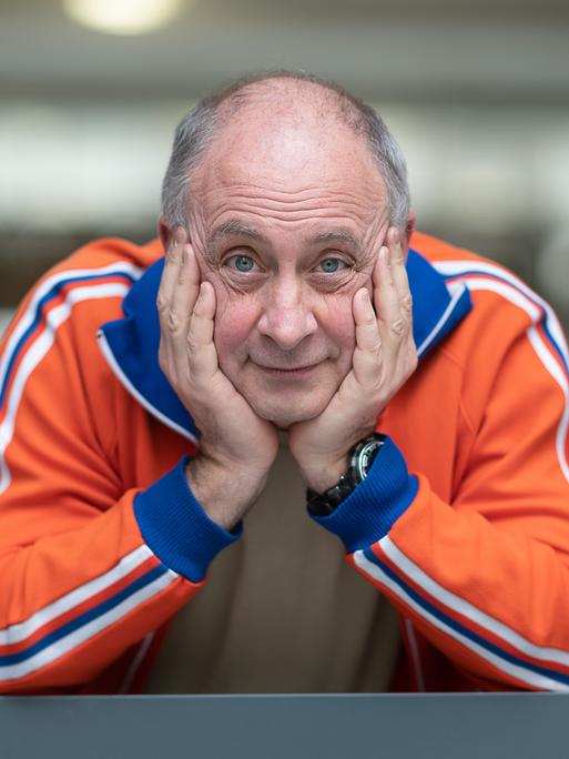 Alfons trägt eine orange-blaue Trainingsjacke und schaut, den Kopf auf die Hände gestützt, freundlich aus dem Fenster direkt in die Kamera.
