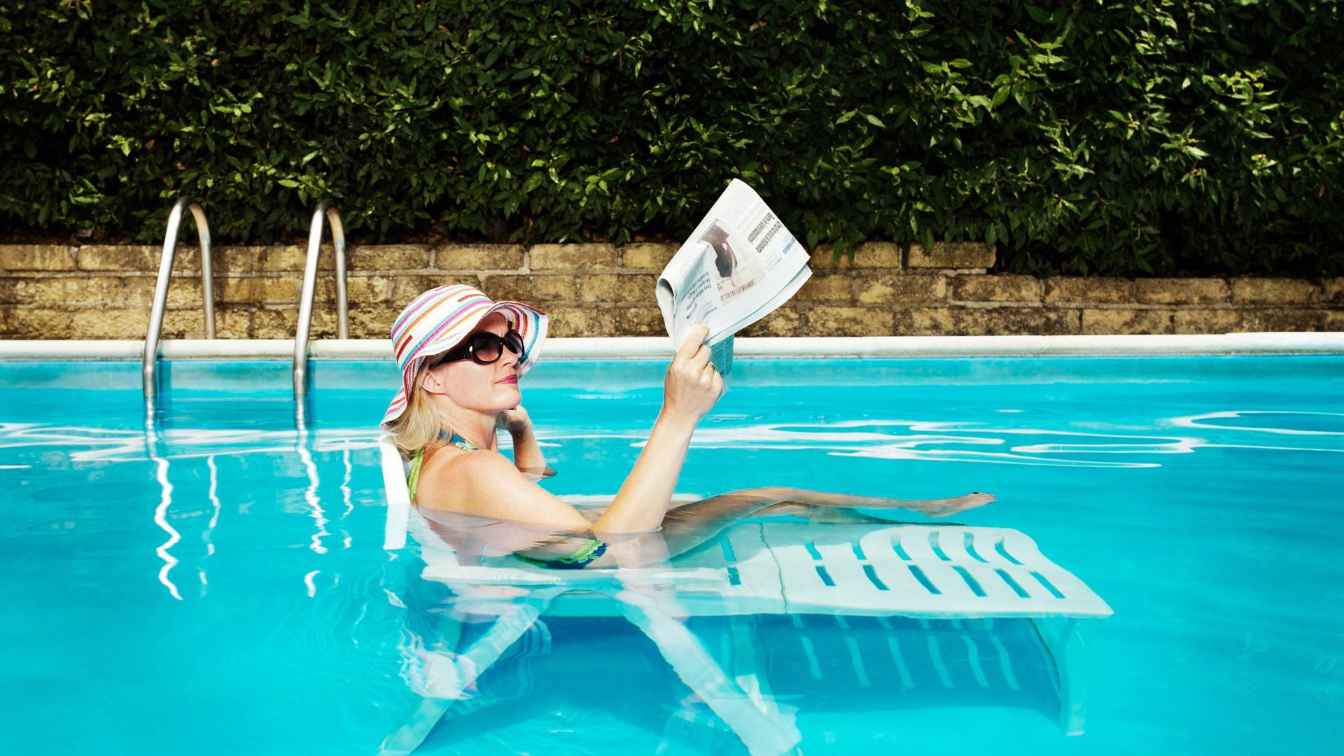 Eine Frau mit einem Sonnenhut liegt auf einer Sonneliege in einem gefüllten Pool und liest Zeitung.